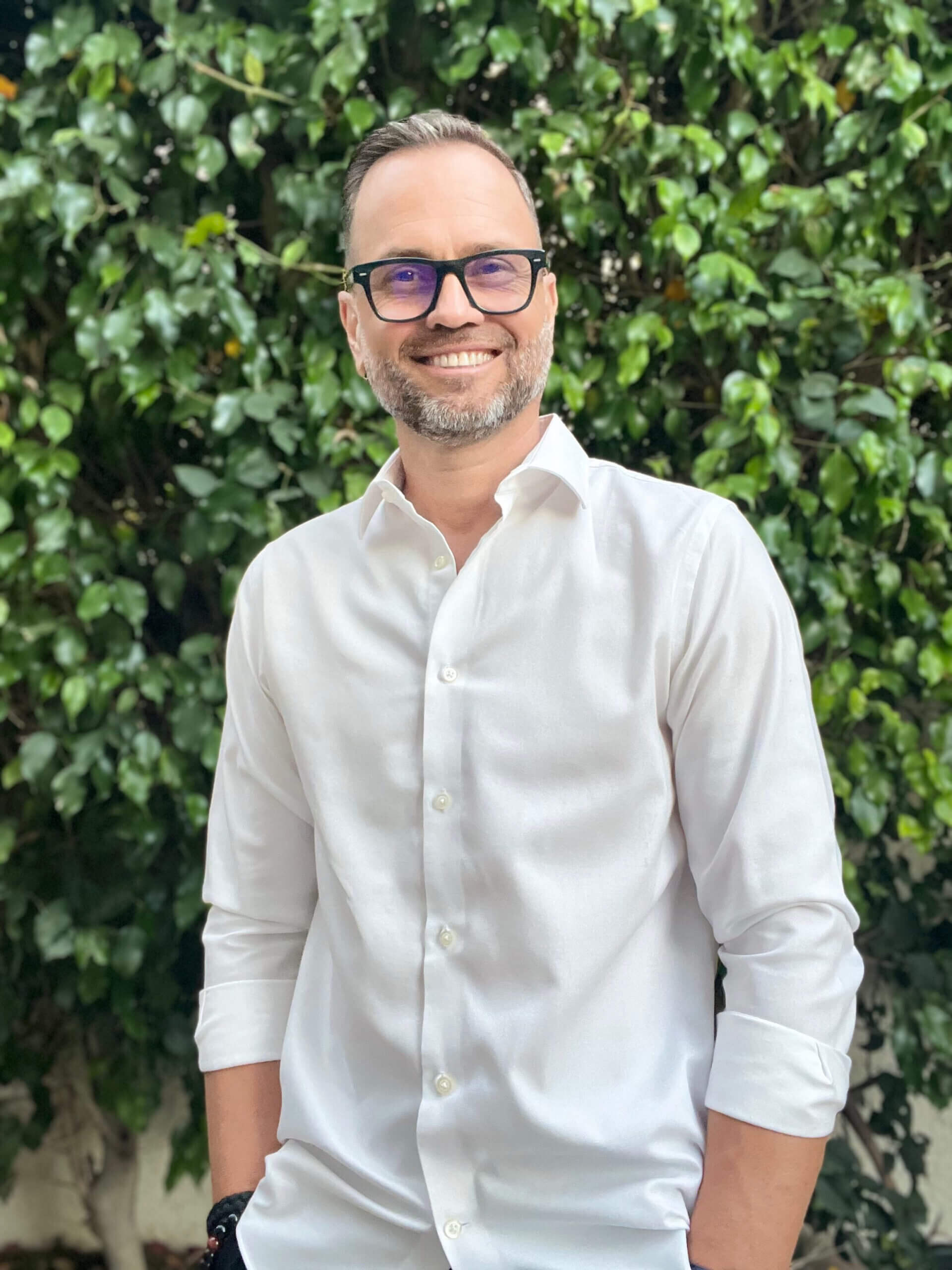 A man in glasses and white shirt standing next to green bushes.